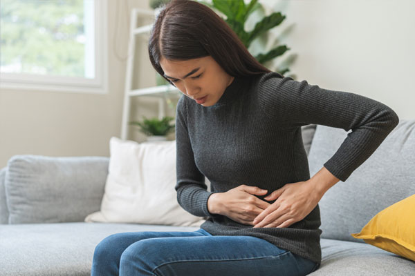 Asian woman sitting on a sofa, holding her lower abdomen with both hands and appearing to experience abdominal discomfort.